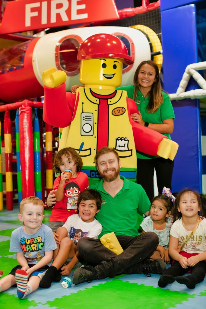 어린이와 어른이 실내 놀이터에서 대형 레고 캐릭터 옆에 앉아 미소를 짓고 있습니다.