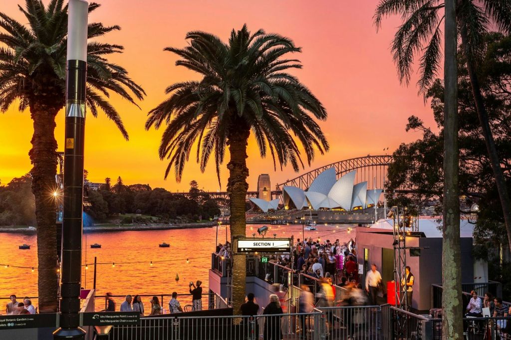 야자수로 둘러싸인 시드니 오페라하우스와 하버 브리지의 활기찬 일몰 전망을 감상할 수 있으며, 물가에 사람들이 모여듭니다.