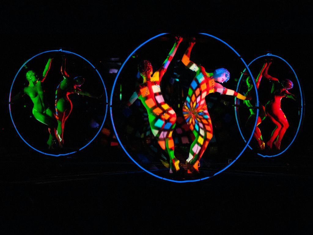 어두운 환경에서 활기찬 조명과 패턴이 있는 원형 프레임 내부의 다채로운 바디 수트를 입은 공연자.