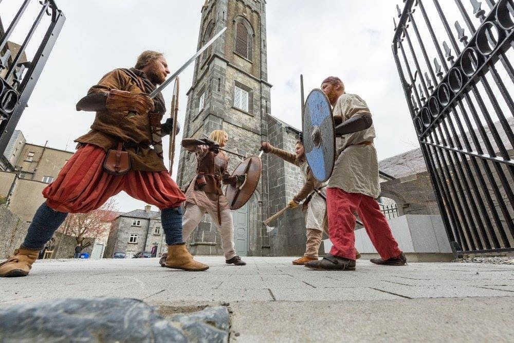 검과 방패를 든 중세 복장을 한 네 명이 탑이 있는 석조 건물 밖에서 모의 전투를 벌입니다.