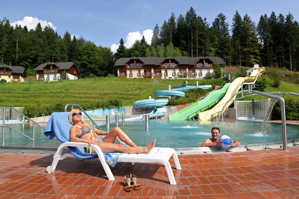 한 사람이 미끄럼틀이 있는 수영장 근처의 라운지 의자에서 휴식을 취하고 있습니다. 물속의 한 남자가 파란 공을 들고 있습니다. 숲과 건물이 배경입니다.