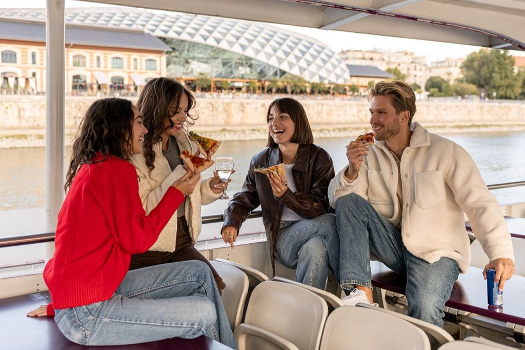도시 풍경과 현대적인 건물을 배경으로 4명이 보트에 앉아 피자를 먹고 대화를 나누고 있습니다.
