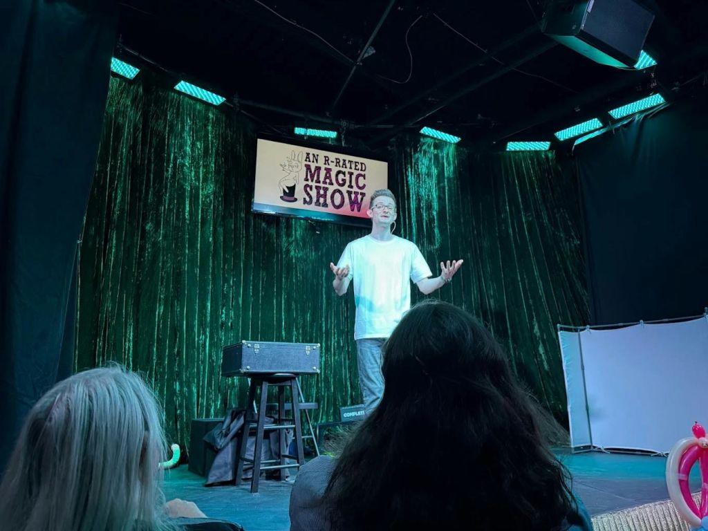 흰색 셔츠를 입은 마술사가 녹색 커튼 배경과 "R 등급의 마술쇼" 라고 적힌 표지판을 들고 무대에서 공연을 합니다.