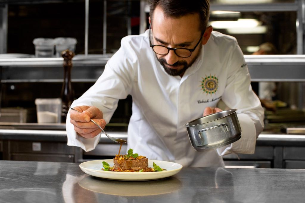 흰색 유니폼을 입은 셰프가 전문 주방에서 소스팬과 숟가락을 들고 정성스럽게 요리를 플레이팅하고 있습니다.