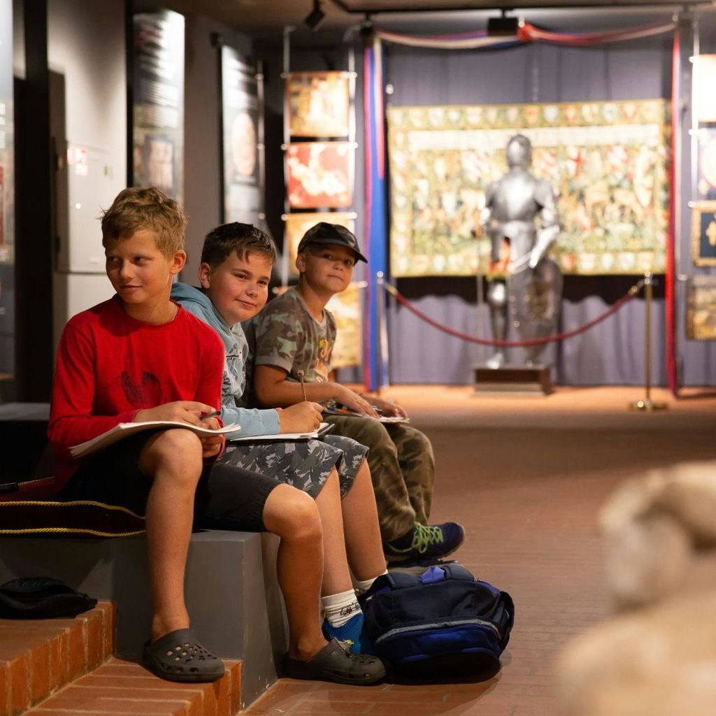 세 명의 소년이 박물관 안 벤치에 앉아 스케치를 하고 있습니다. 그 뒤에는 갑옷을 포함한 역사적인 전시가 있습니다.