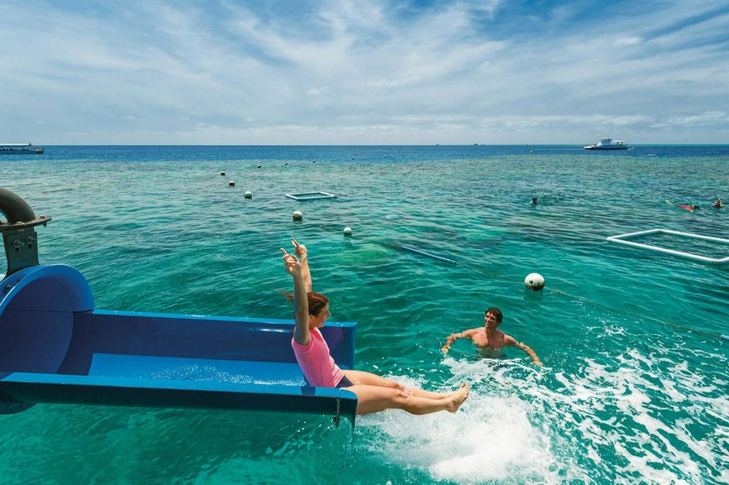 한 사람이 푸른 미끄럼틀에서 바다로 미끄러져 들어가고, 근처의 다른 사람이 물속에 있습니다. 하늘이 부분적으로 흐리다.