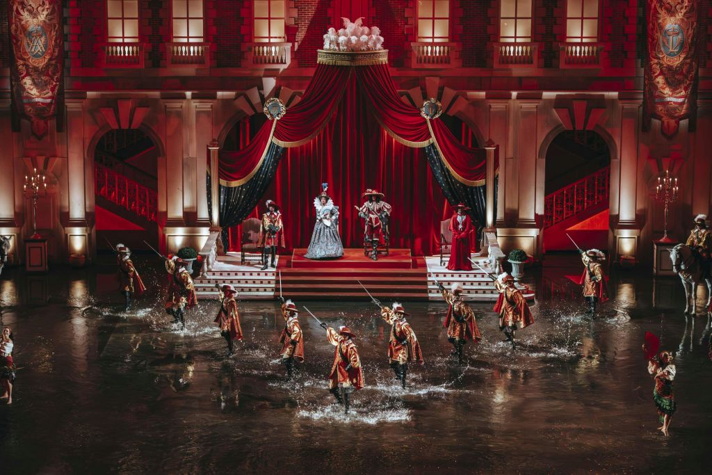 왕실을 배경으로 물로 가득 찬 무대에서 공연하는 기간 의상을 입은 배우들의 연극 공연입니다.