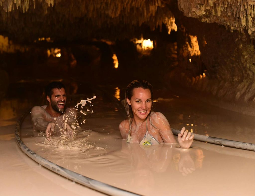 수영복을 입은 두 사람이 천장에 종유석이 매달린 희미한 조명의 동굴에서 진흙 목욕을 즐기고 있습니다.