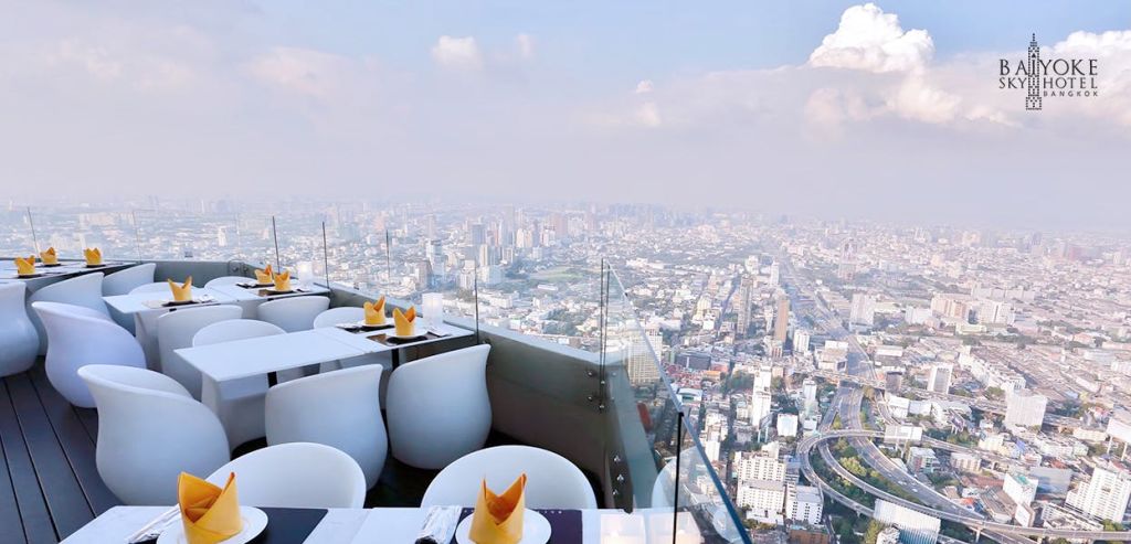 흰색 의자, 냅킨이 세팅된 테이블, 유리 장벽이 있는 야외 옥상 레스토랑에서 도시 스카이라인의 전경을 감상하실 수 있습니다.