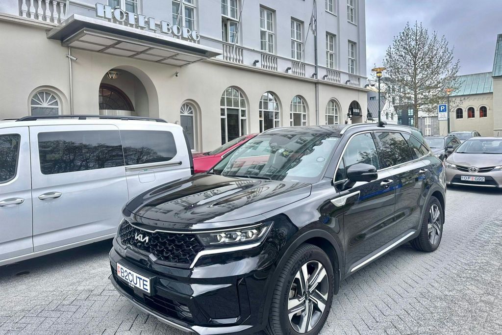 맞춤형 "B2CUTE" 번호판을 갖춘 검은색 기아 SUV가 호텔 보그 앞에 주차되어 있습니다. 근처에는 흰색 밴과 빨간색 자동차가 있습니다.