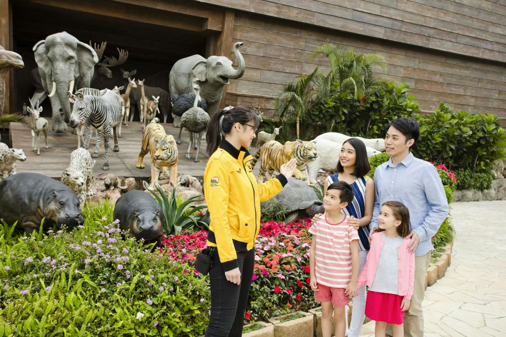 노란색 재킷을 입은 가이드가 어트랙션 밖에 있는 실물 크기의 동물 조각상 네 개로 구성된 웃는 가족과 이야기를 나누고 있습니다.