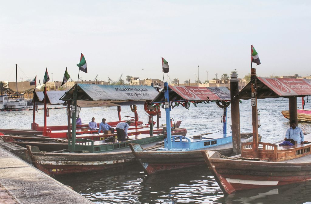 차양과 깃발이 달린 작고 화려한 보트가 물가에 정박해 있습니다. 세 사람이 파란색 보트에 탑승합니다. 건물과 나무를 배경으로.