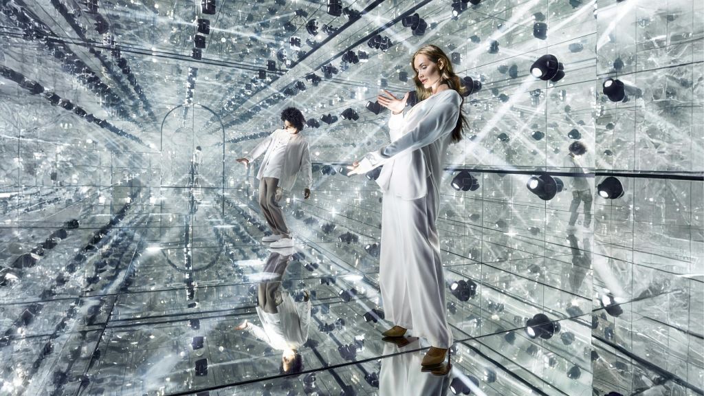 흰색 의상을 입은 두 사람이 거울로 된 벽과 바닥이 있는 방에서 포즈를 취하며 여러 번 반사되는 만화경 효과를 연출합니다.