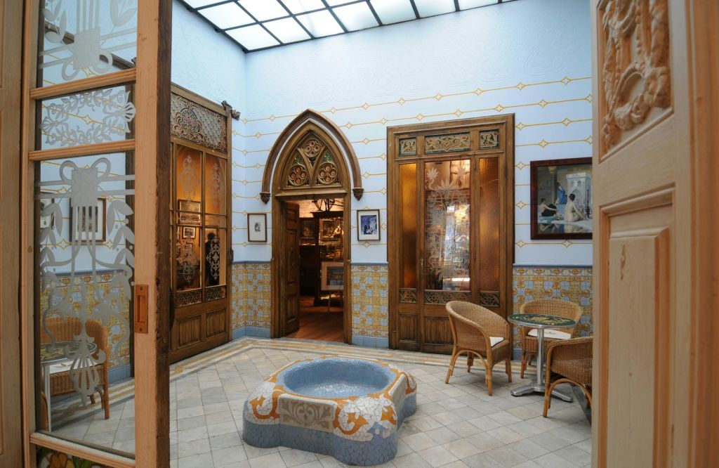 나무 문, 타일 벽, 고리 버들 가구, 둥근 돌 분수, 유리 천장이 있는 화려한 실내 안뜰.