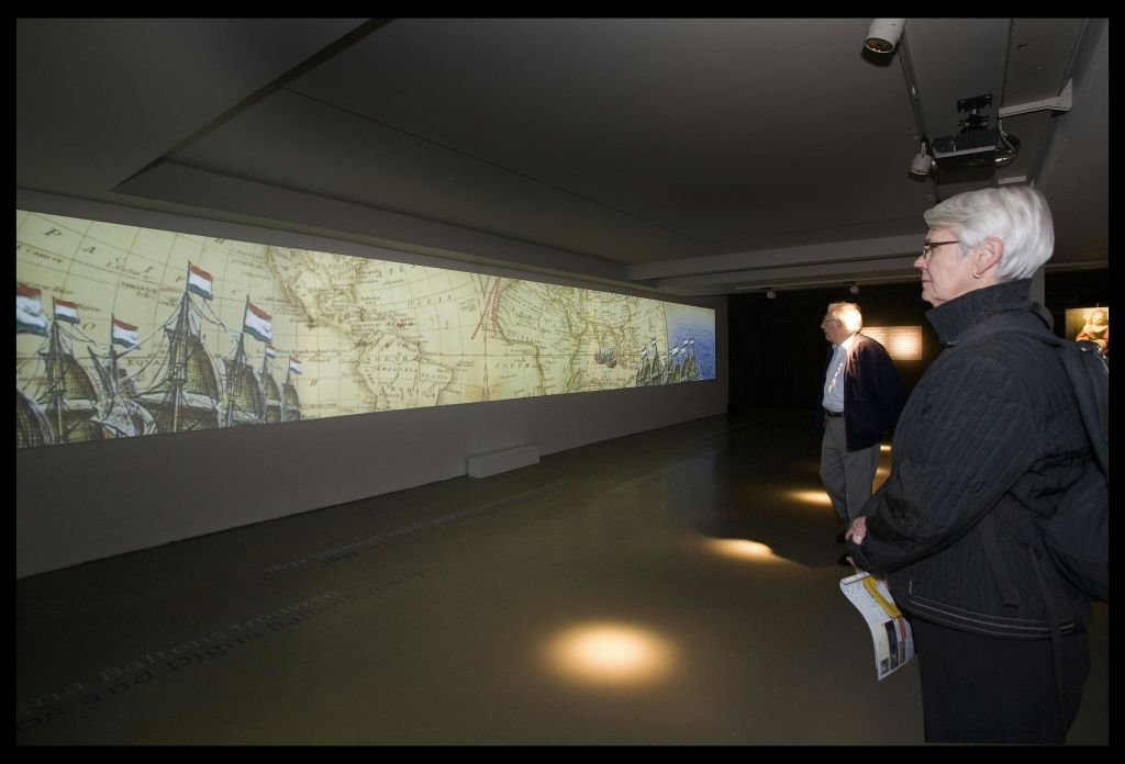 벽에 투영된 커다란 역사 지도를 바라보는 조명이 희미한 전시실에 앉아 있는 두 사람.