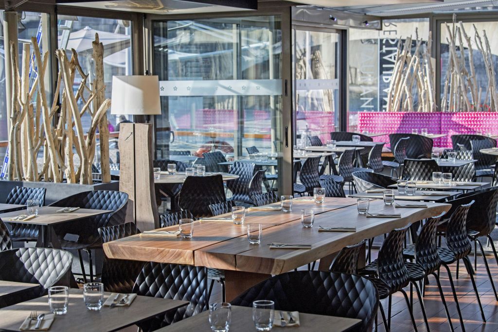 커다란 창문 옆에 나무 테이블, 검은색 퀼트 의자, 투명한 유리잔, 식기가 깔끔하게 배치된 현대적인 레스토랑.