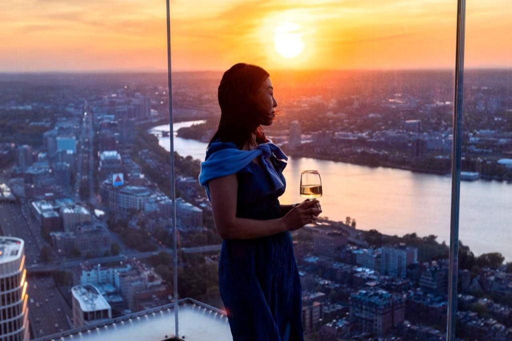 푸른 드레스를 입은 한 여성이 높은 건물에서 석양을 감상하며 도시 풍경과 강을 바라보며 와인잔을 들고 있습니다.