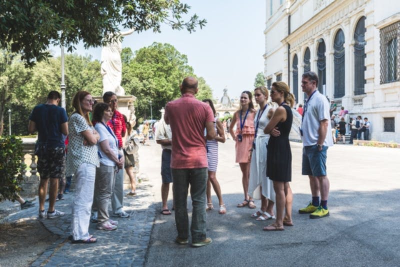 화창한 날 화려하게 꾸며진 커다란 건물 밖에서 가이드의 말을 들으며 서 있는 사람들의 무리. 배경에는 나무와 조각상이 보입니다.