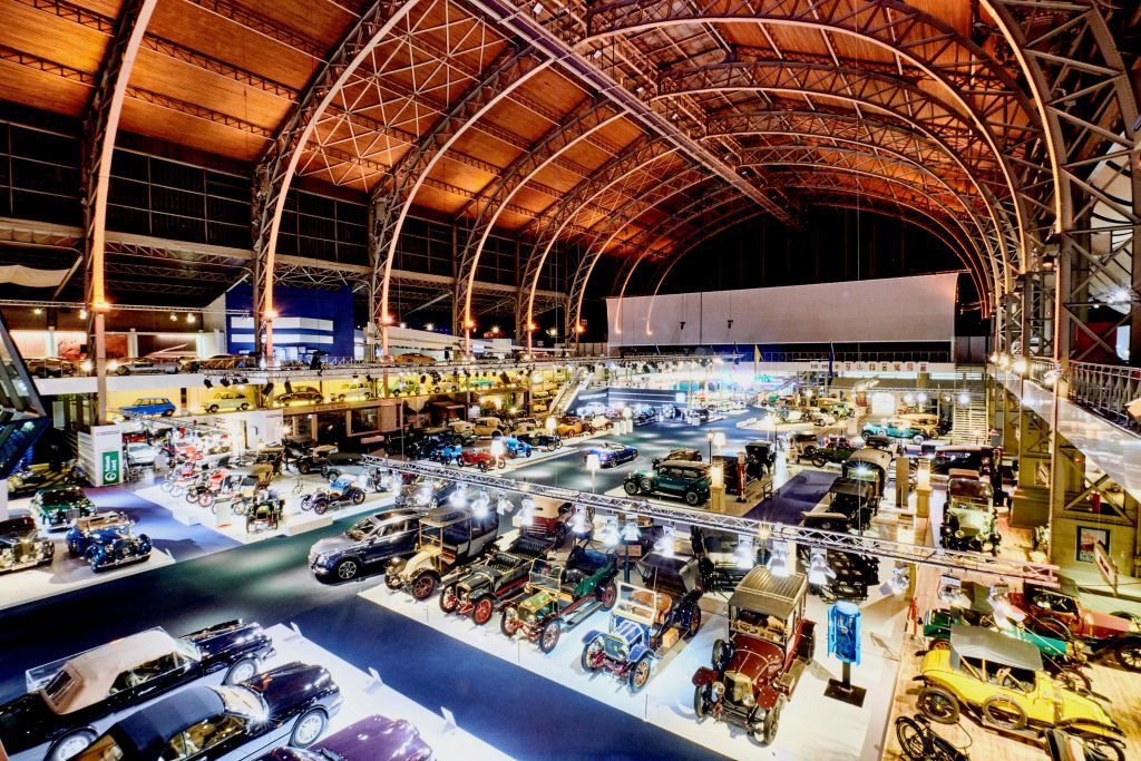 밝은 조명이 있는 높고 아치형 천장 아래 빈티지 자동차와 클래식 자동차를 전시한 대형 실내 자동차 박물관입니다.