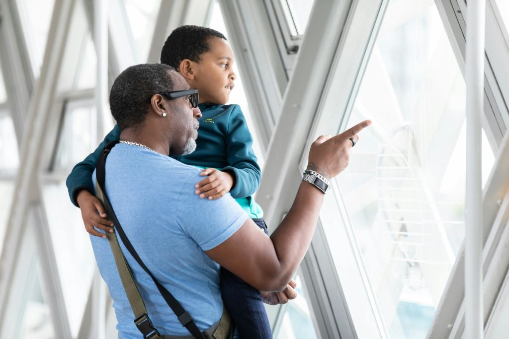 아이를 안고 있는 어른이 커다란 창밖을 내다보고 있습니다. 이 장면은 기하학적인 디자인이 돋보이는 현대적인 건물에 있는 것 같습니다.