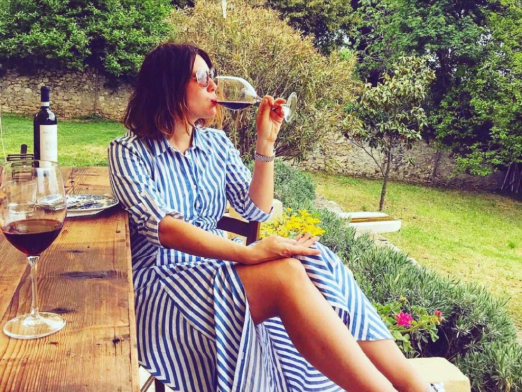 줄무늬 드레스를 입은 여성이 녹음으로 둘러싸인 야외 나무 테이블에서 와인 병과 유리잔을 들고 와인을 음미하고 있습니다.