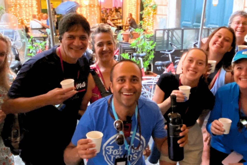 한 무리의 사람들이 음료를 들고 매달린 조명과 녹지가 있는 야외 카페 앞에서 포즈를 취하고 있습니다.