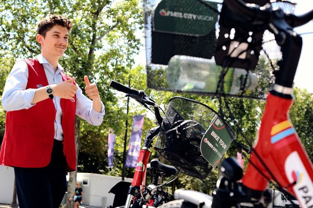 나무와 현수막을 배경으로 한 야외 공원에서 빨간 조끼를 입은 사람이 자전거 옆에서 엄지손가락을 치켜세우고 있습니다.