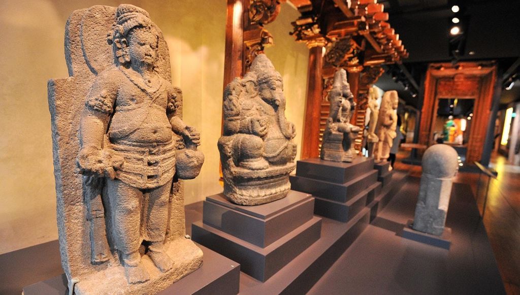 박물관의 받침대에 고대 석상이 줄지어 있고, 배경에는 정교하게 조각된 나무 기둥이 있습니다.