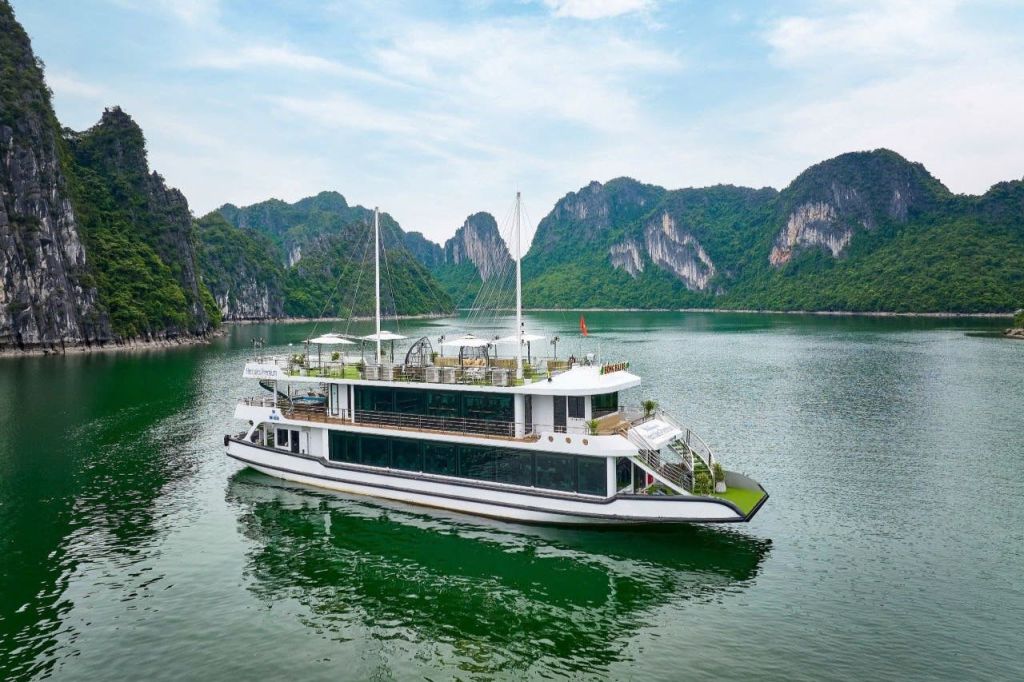 부분적으로 흐린 하늘 아래 푸른 언덕과 석회암 절벽을 배경으로 잔잔한 물 위에 하얀 멀티 데크 보트가 떠 있습니다.