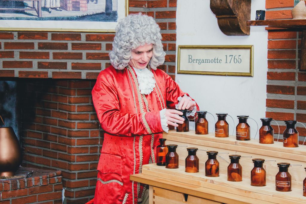 흰 가발을 쓴 빨간색 역사 의상을 입은 사람이 벽돌 벽 근처의 나무 테이블에서 호박색 병을 살펴보고 있습니다.