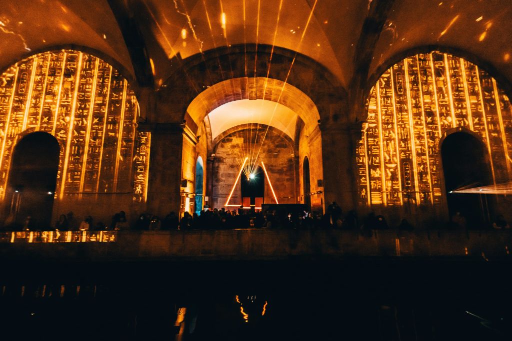 중앙에 석조 아치, 조명 상형문자 패널, 삼각형 조명 디스플레이가 있는 조명이 희미한 방입니다. 실루엣의 사람들.