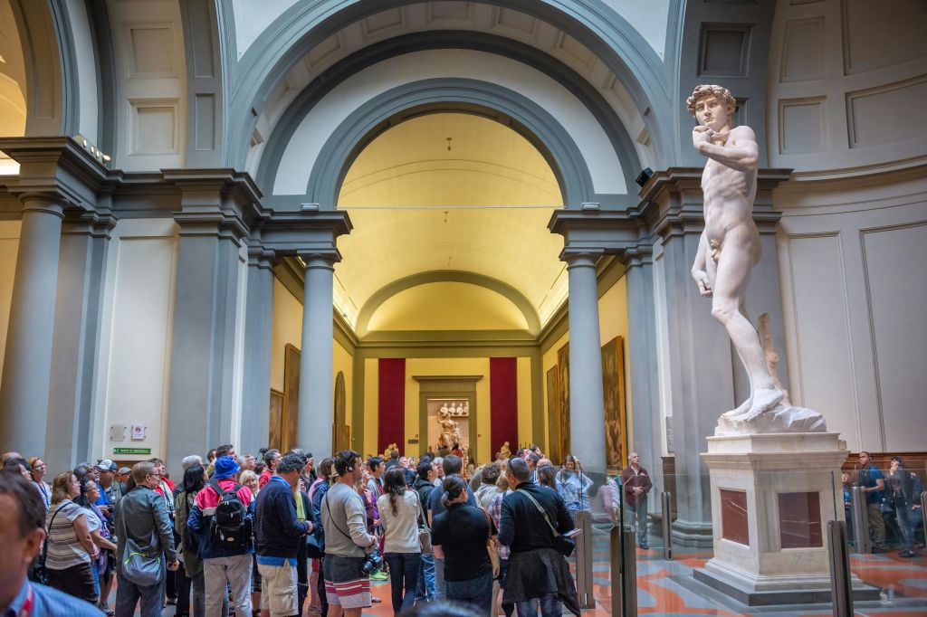 오른쪽에는 남성 인물의 커다란 동상이 있고 배경에는 아치형 천장이 있는 박물관 홀에는 수많은 관람객이 모여 있습니다.