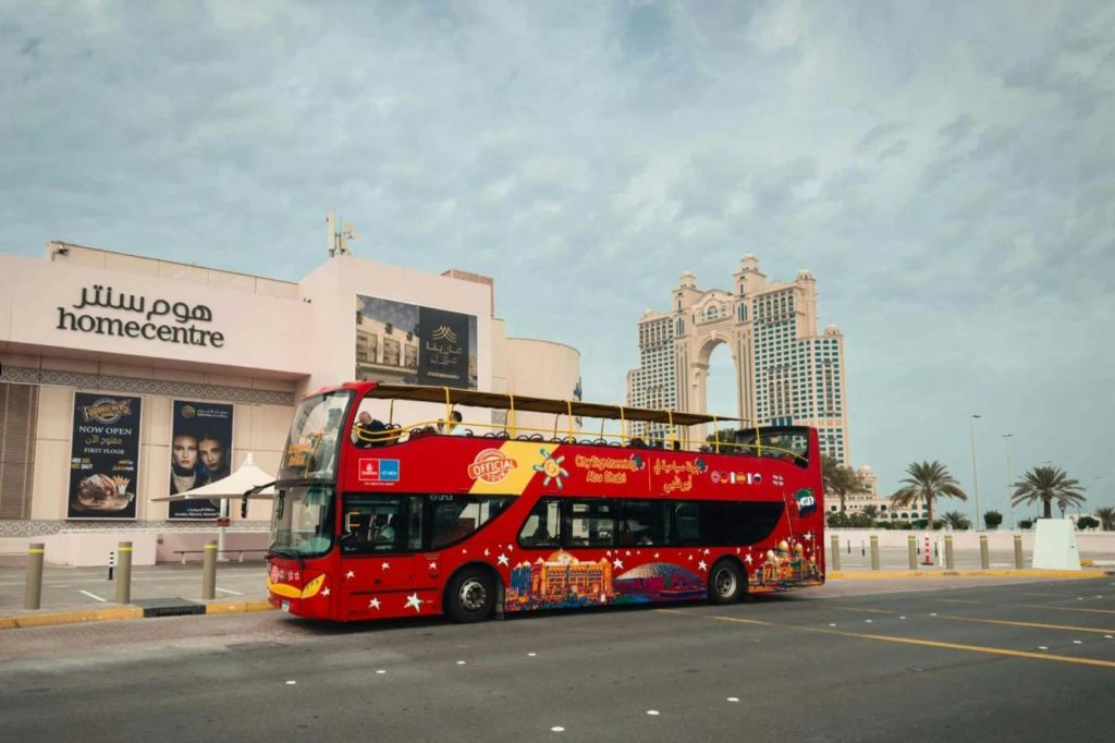 건물과 야자수를 배경으로 한 거리에 빨간색 2층 관광버스가 주차되어 있습니다.