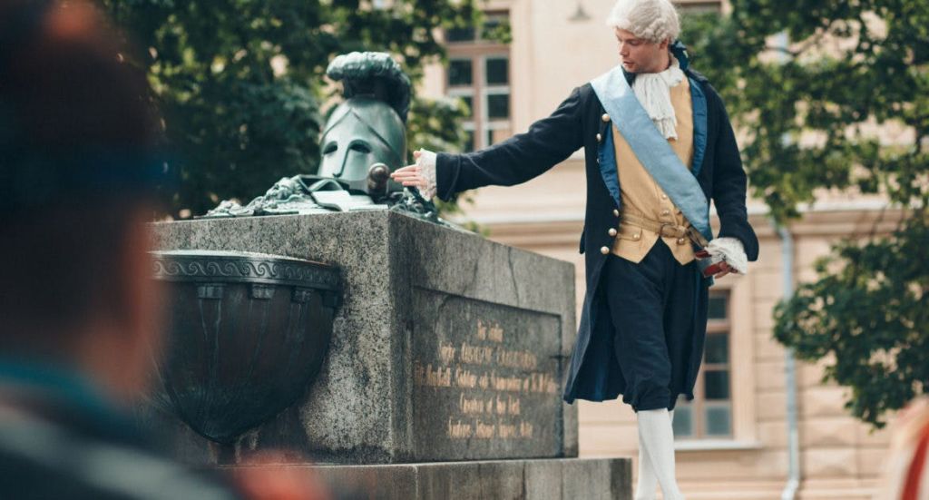 파란색 띠와 삼각 모자가 달린 역사적인 복장을 한 남성이 돌 받침대 옆에 서 있습니다. 나무와 건물이 배경입니다.