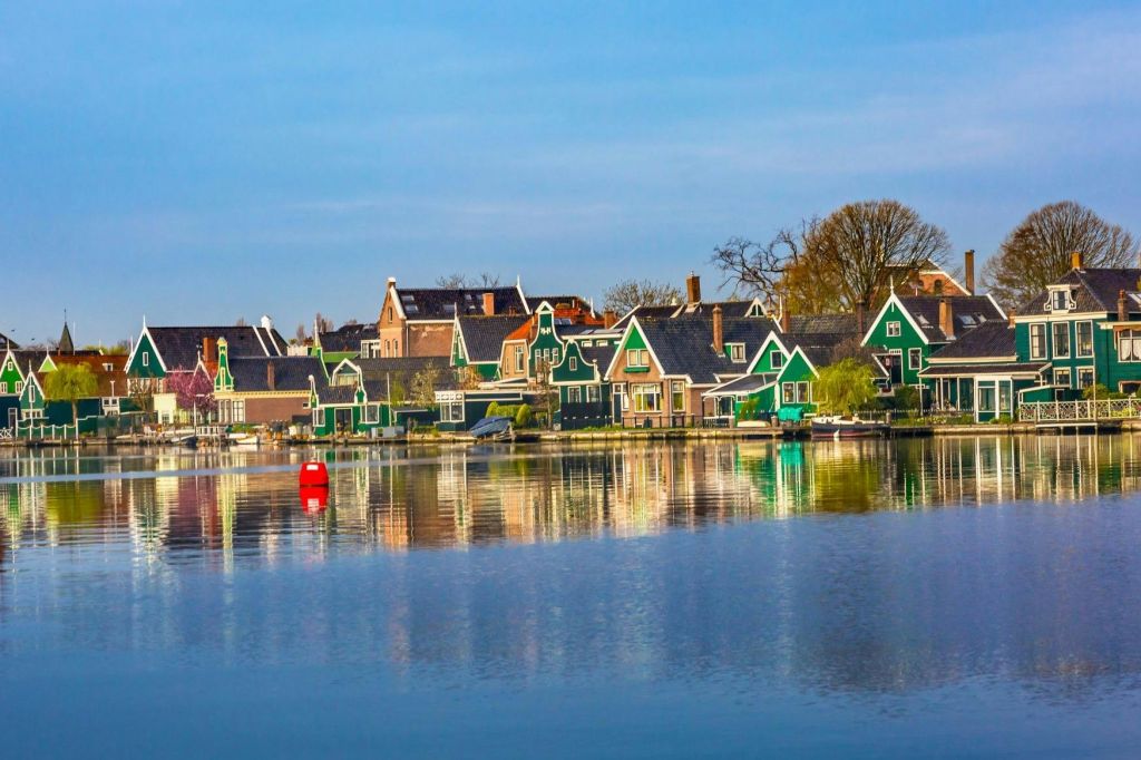 조용한 바닷가에 가파른 지붕이 있는 다채로운 주택이 있습니다. 푸른 하늘 아래에는 나무와 밝은 붉은 부표도 보입니다.