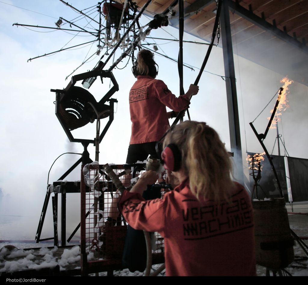 매칭되는 레드 재킷을 입은 두 사람은 다양한 레버와 불타는 불꽃으로 복잡하고 기계적인 설정을 작동합니다.