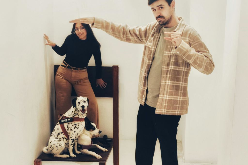 격자무늬 셔츠를 입은 남성이 벽에 기대어 있는 여성 옆에서 두 마리의 개가 나무 벤치에 함께 앉아 있는 제스처를 취하고 있습니다.