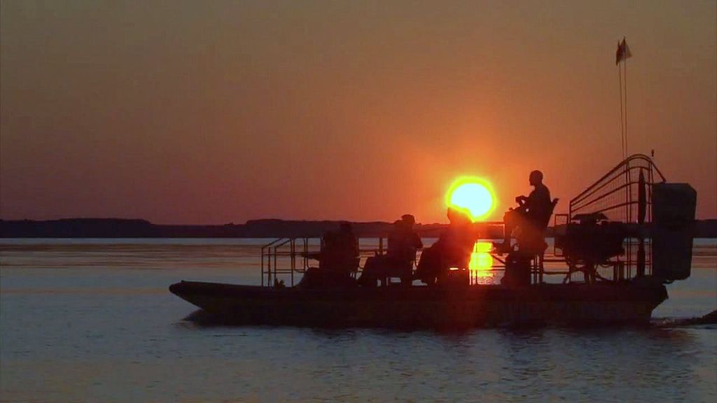 실루엣의 사람들은 잔잔한 물 위로 지는 석양에 보트에 앉아 있습니다. 태양은 수평선 근처에서 주황색 빛을 발합니다.