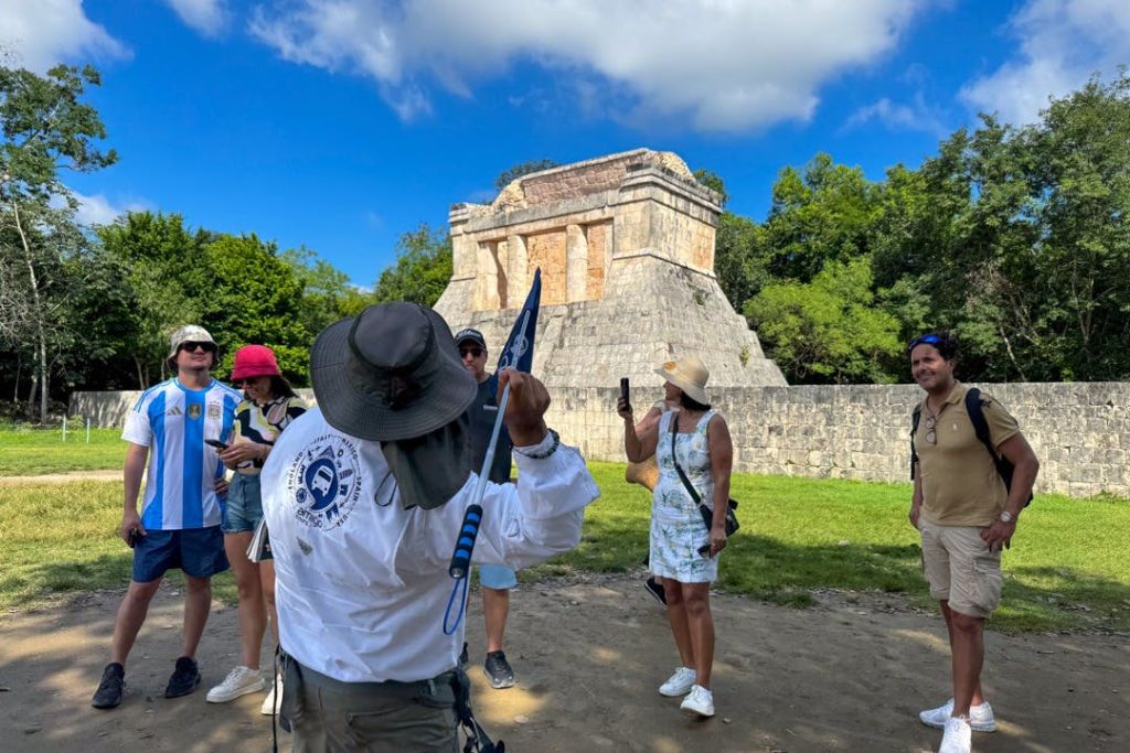 세 사람이 녹지로 둘러싸인 고대 석조 건물 근처에 서 있으며, 한 명은 깃발을 들고 있고 다른 한 명은 전화기를 사용하고 있습니다.