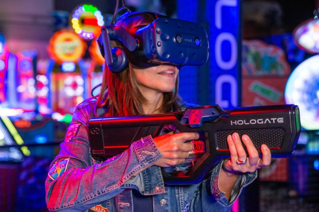 다채로운 조명을 배경으로 VR 헤드셋을 착용하고 아케이드 환경에서 게이밍 건 컨트롤러를 들고 있는 사람.