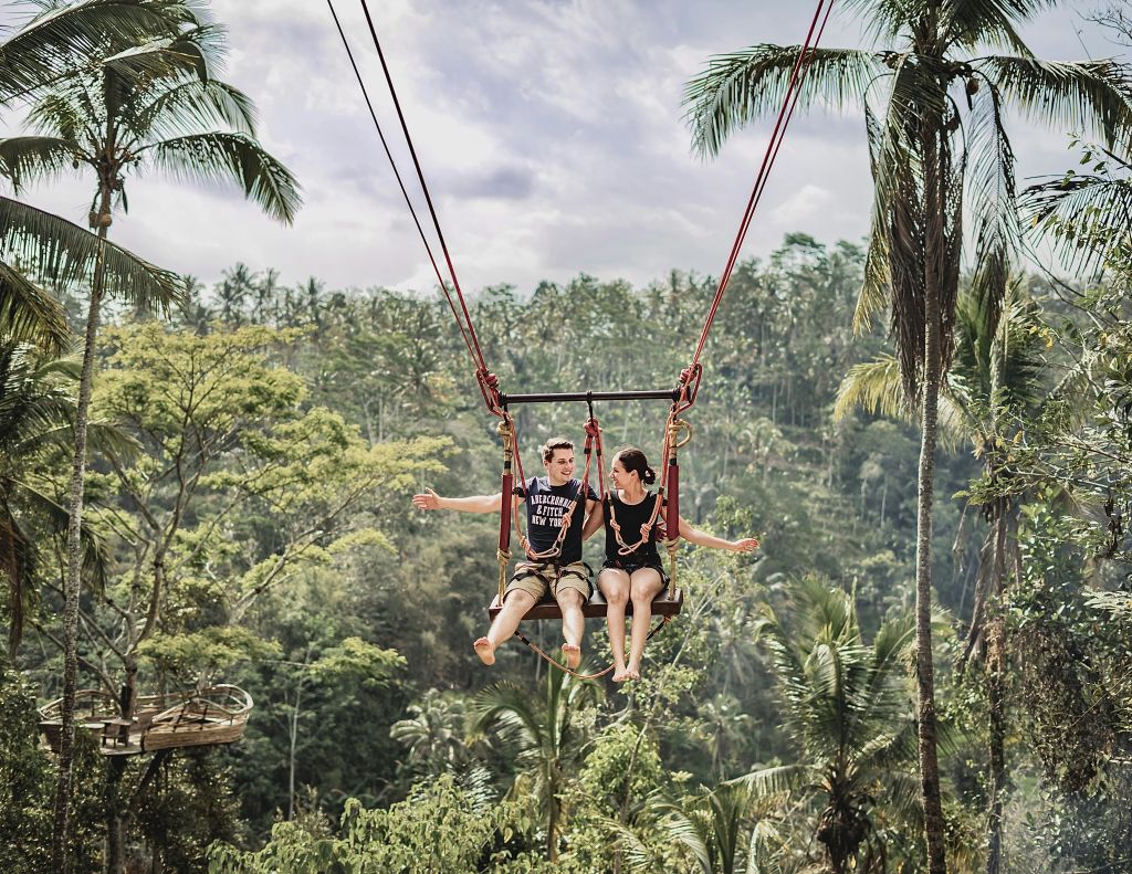 한 남녀가 야자수를 배경으로 울창한 열대 숲 위에 높이 매달린 그네에 앉아 있습니다.