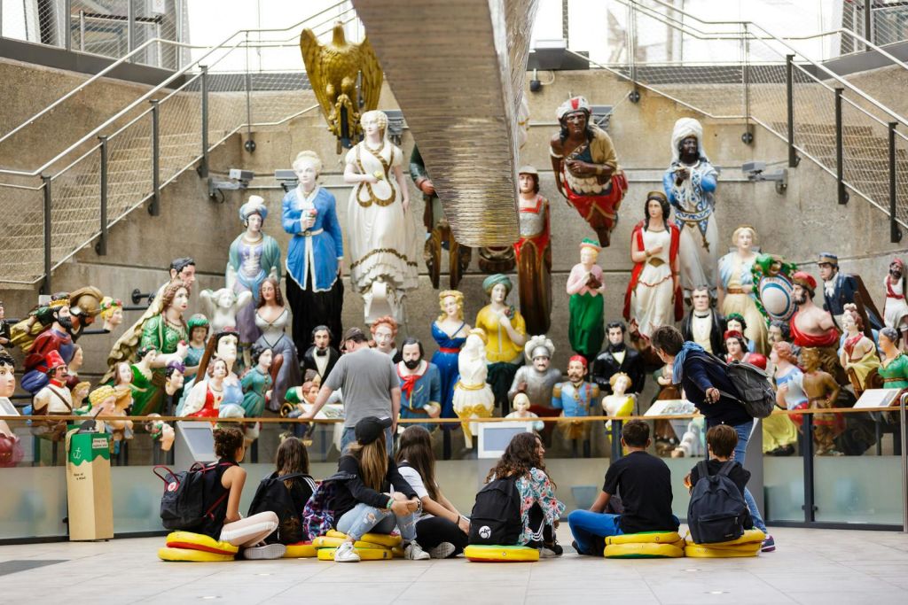 한 무리의 사람들이 쿠션에 앉아 유리 장벽 뒤에 전시된 역사 및 문화 동상 전시를 관찰하고 있습니다.