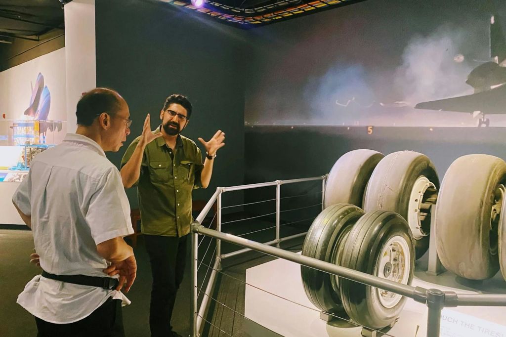 두 남자가 전시 공간에서 대화를 나누고 있는데, 한 남자가 제스처를 취하고 있습니다. 산업용 물체가 전경에 부분적으로 보입니다.