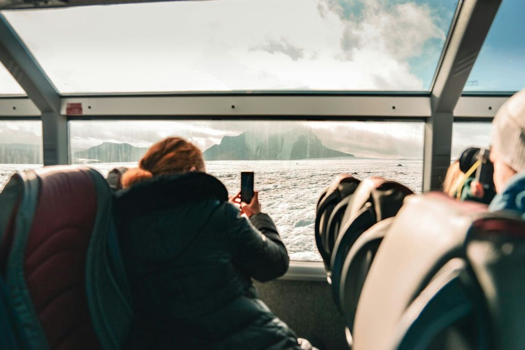 눈 덮인 풍경과 산이 내려다보이는 커다란 창문이 있는 버스 같은 차량 안에서 휴대폰으로 사진을 찍는 사람.