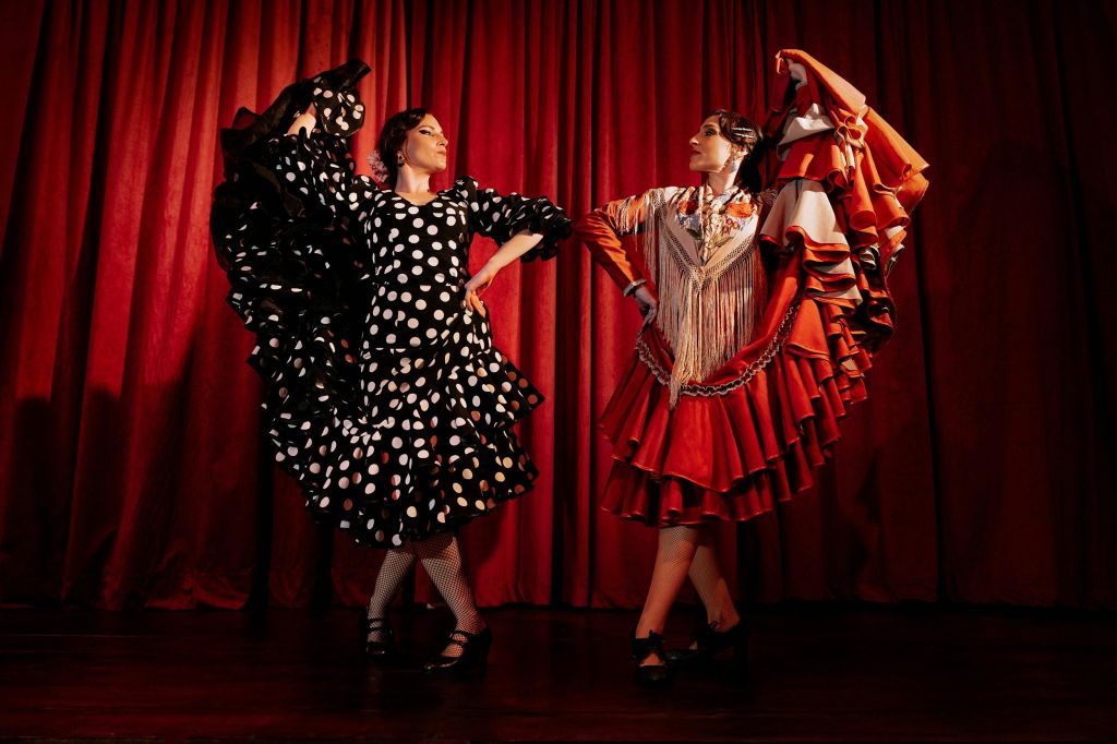 빨간색 커튼 앞에서 두 명의 여성이 전통 드레스를 입고 플라멩코 춤을 추고 있습니다. 하나는 물방울 무늬가 있는 검은색, 다른 하나는 프린지가 있는 빨간색입니다.