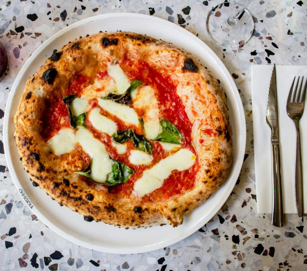 테라조 테이블의 포크와 나이프 옆에 흰색 접시에 모짜렐라, 토마토 소스, 바질을 올린 나폴리식 피자.