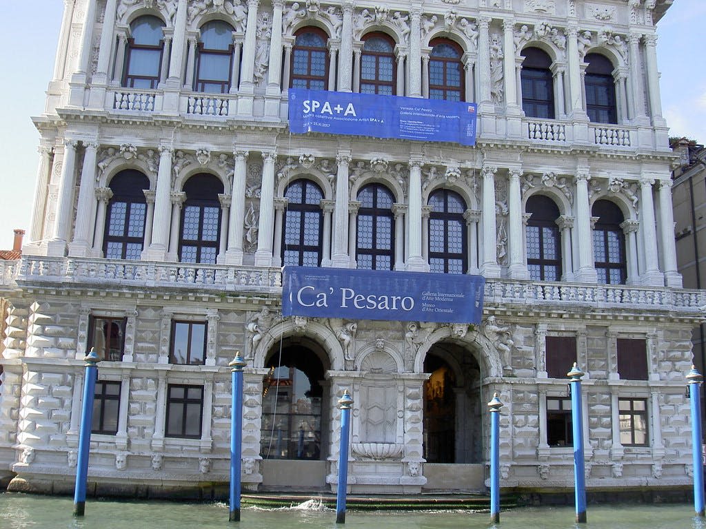 아래 운하에 두 개의 파란색 배너와 파란색 기둥이 있는 베네치아의 카페사로 (Ca 'Pesaro) 의 화려한 외관.