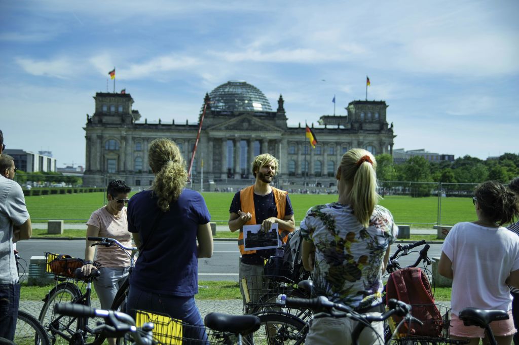 주황색 조끼를 입은 투어 가이드가 유리 돔이 있는 유서 깊은 대형 건물 앞에서 자전거를 타고 세 사람에게 말을 건넨다.