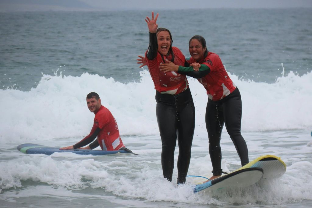 빨간색 셔츠를 입은 두 명의 여성이 서핑보드에 서서 미소를 짓고 손을 흔들고 있으며, 빨간색 셔츠를 입은 한 남성이 서핑보드 배경에 앉아 있습니다.