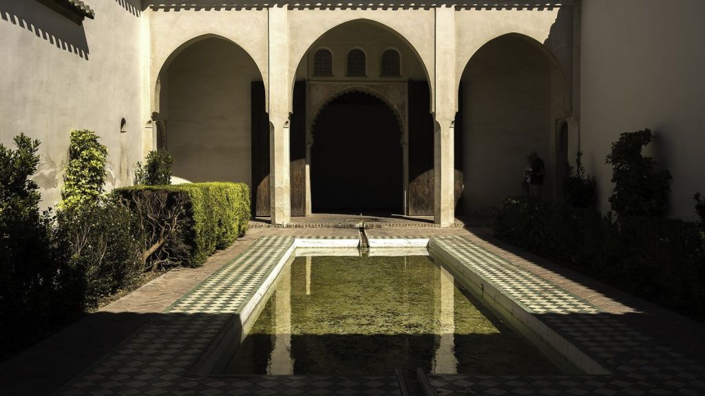 직사각형의 반사 수영장, 타일 바닥, 아치형 출입구, 벽을 따라 다듬어진 녹지가 있는 안뜰입니다.